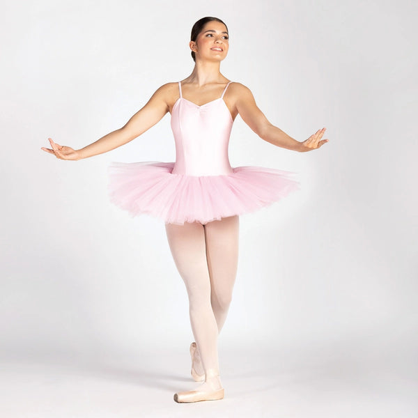 A ballerina posing in a light pink sleeveless leotard with a matching pink tulle tutu and pink tights, set against a plain light gray studio background.