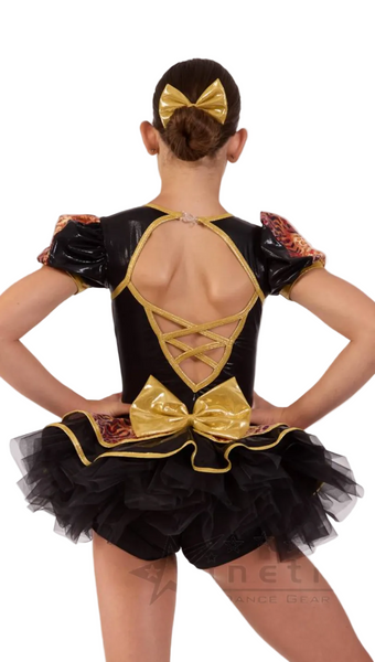 A child model shown from the back wearing a black and gold ballet or dance costume with a large bow at the waist, crisscross back straps, sheer patterned puff sleeves, and a black tutu skirt.