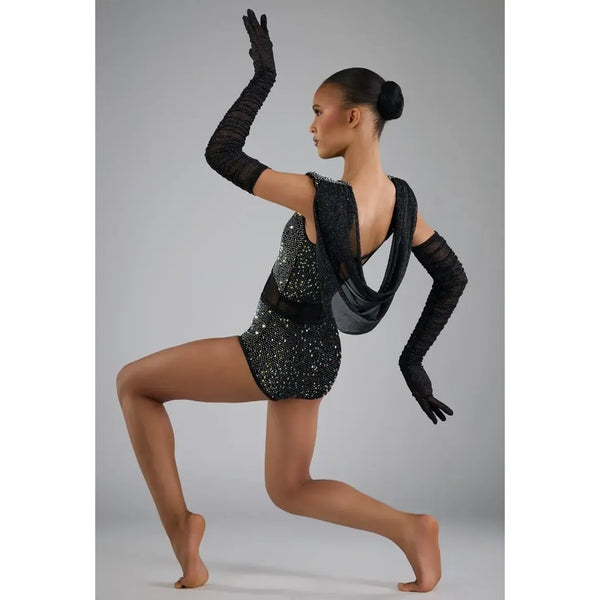 A female dancer in a black sequined leotard with long black gloves, captured in a dynamic lunge pose against a neutral gray studio background.